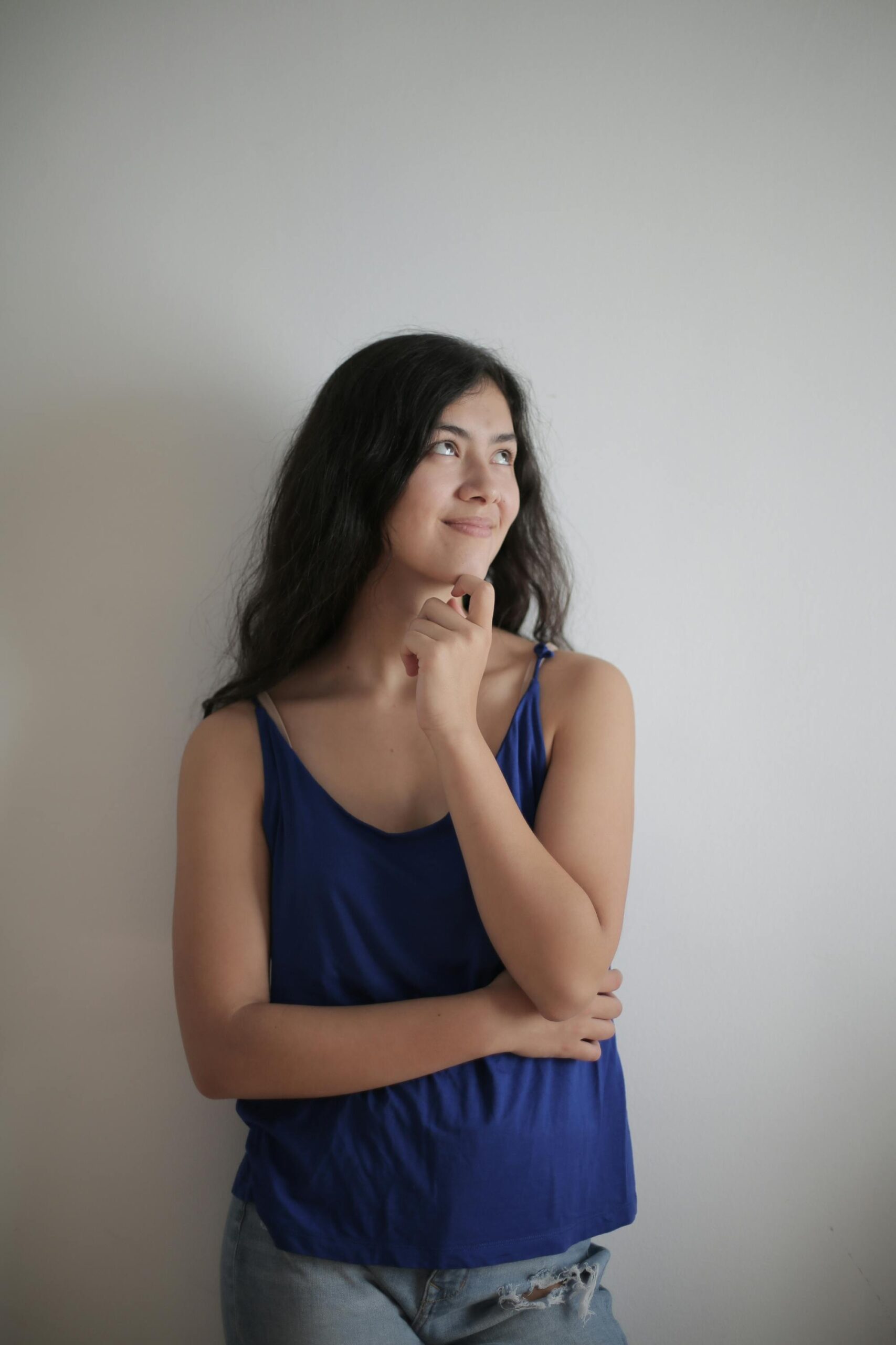 A pensive woman in a blue tank top stands against a plain white wall, smiling gently.